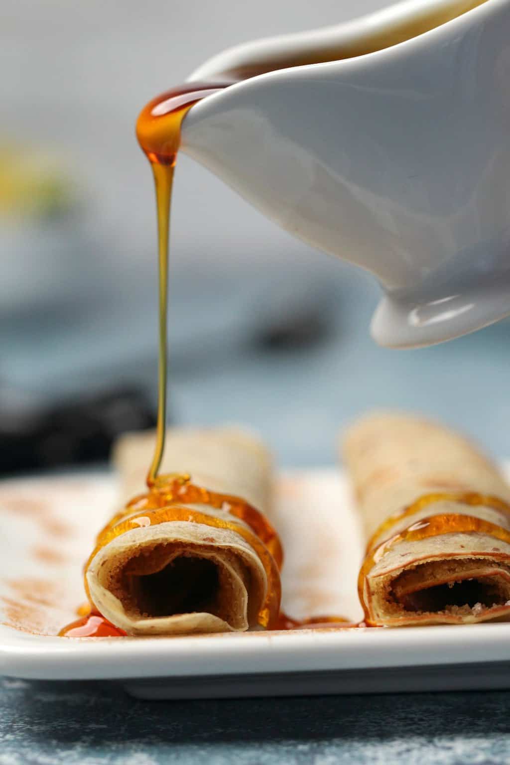 Syrup drizzling over rolled vegan crepes on a white plate. 