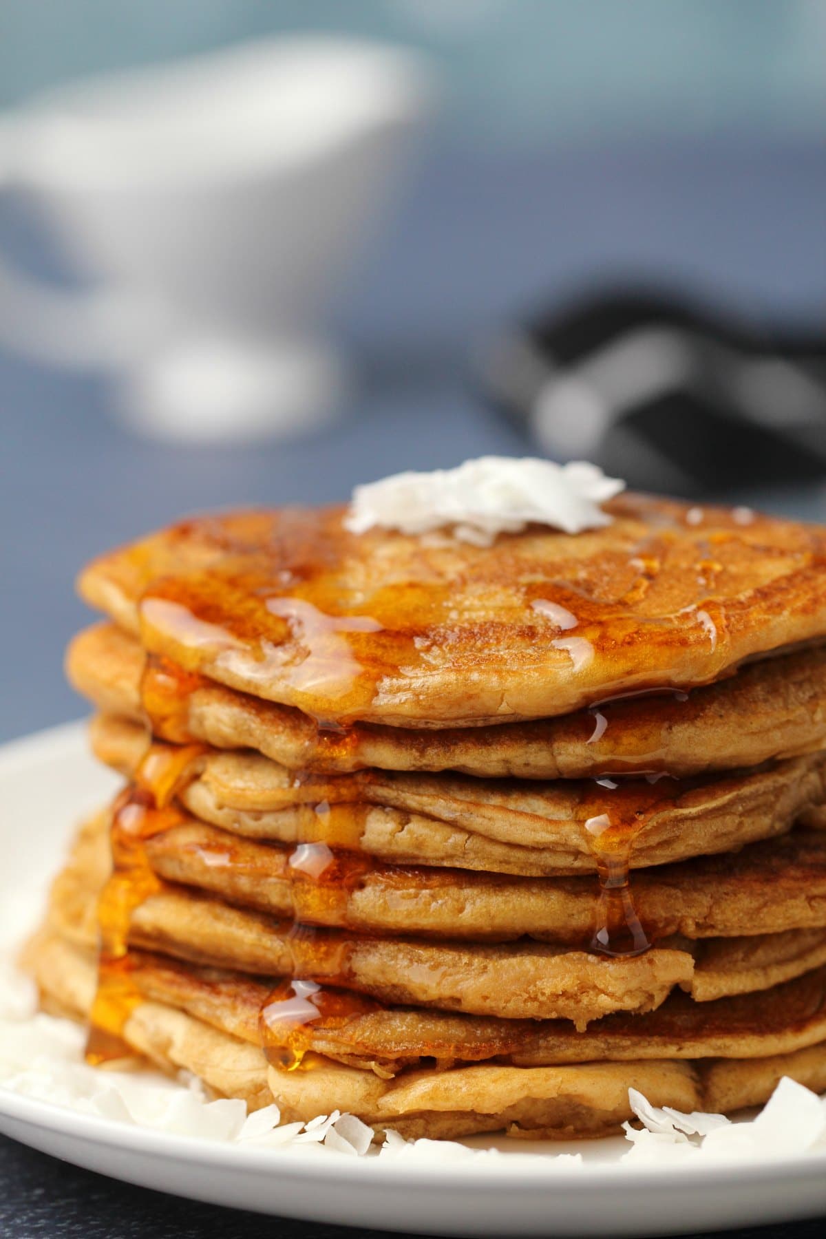 Vegan coconut pancakes topped with coconut flakes and syrup stacked up on a white plate. 