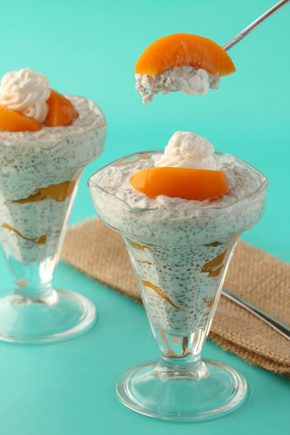 A spoonful of chia pudding hovering above a sundae glass layered with chia pudding and sliced peaches and topped with vegan whipped cream. 