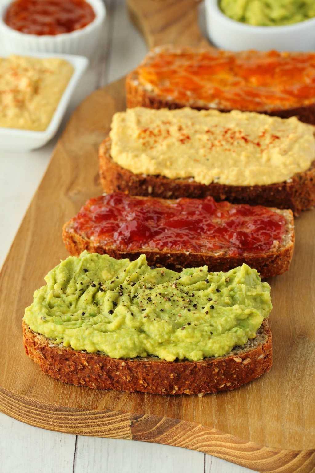 Whole wheat bread slices topped with avocado, jams and hummus on a wooden board. 