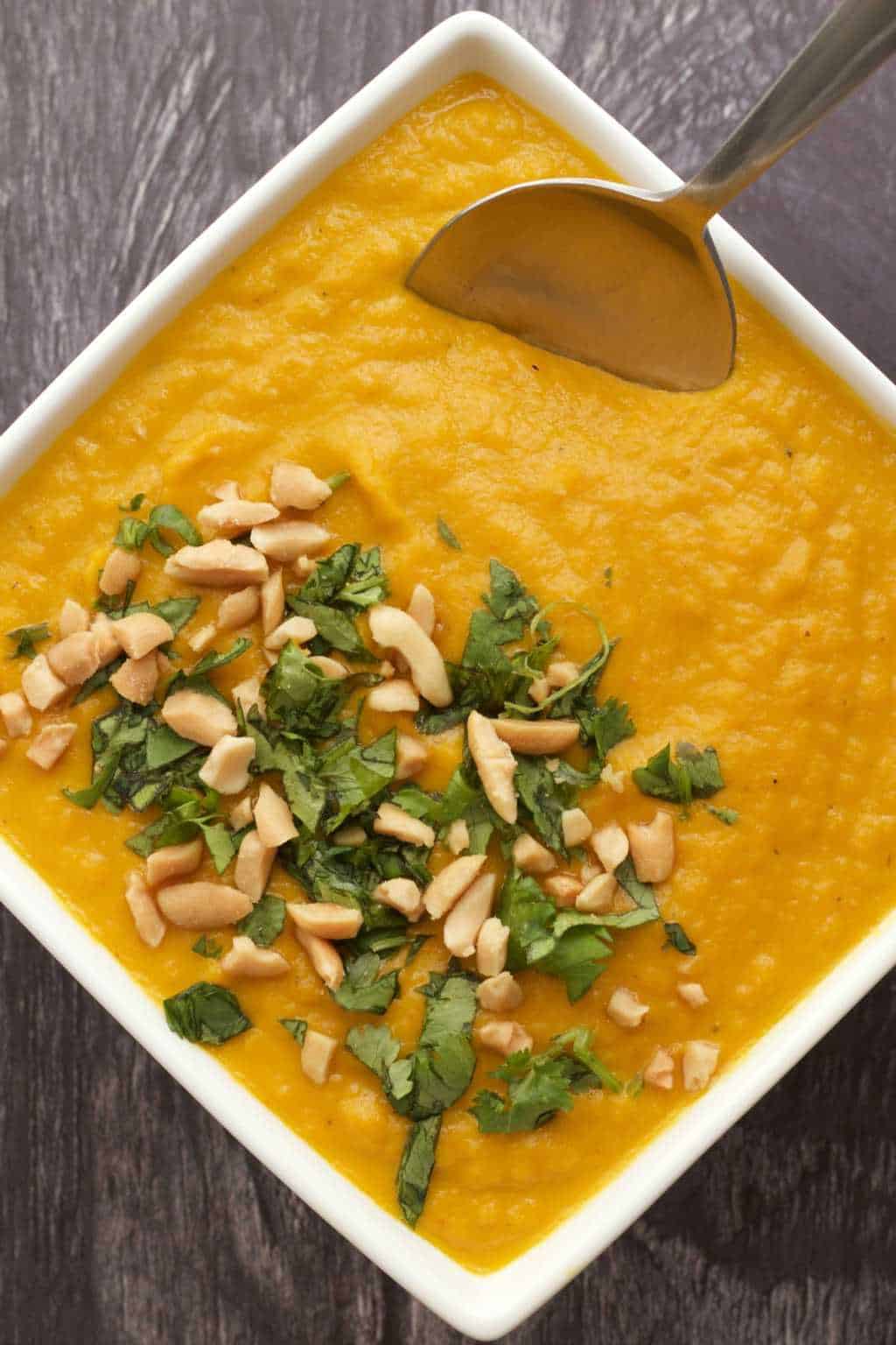 Vegan carrot soup topped with peanuts and chopped cilantro in a white bowl with a spoon. 