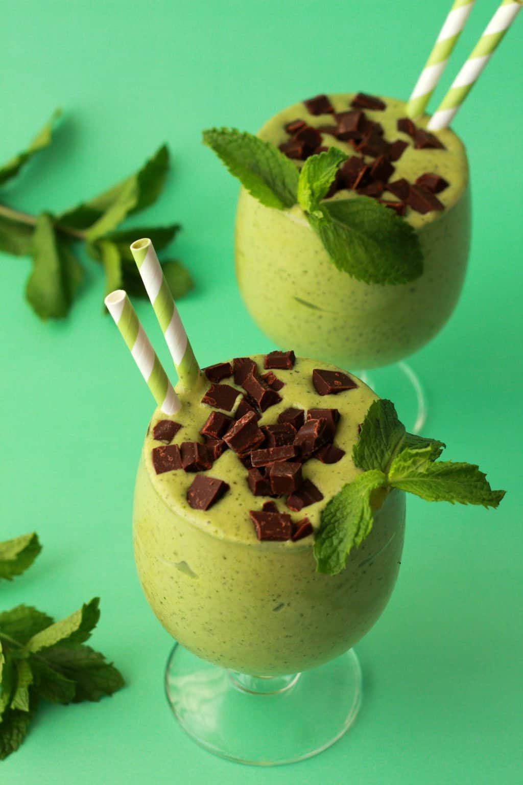 Mint smoothie topped with chopped chocolate in a glass with a green and white striped straw. 