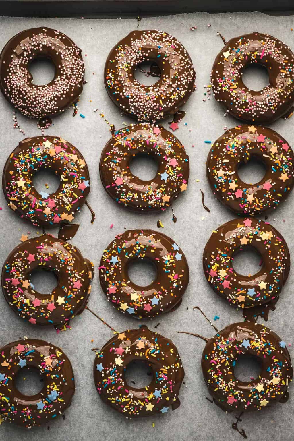 Vegan chocolate donuts topped with melted chocolate and sprinkles on a parchment lined baking tray. 