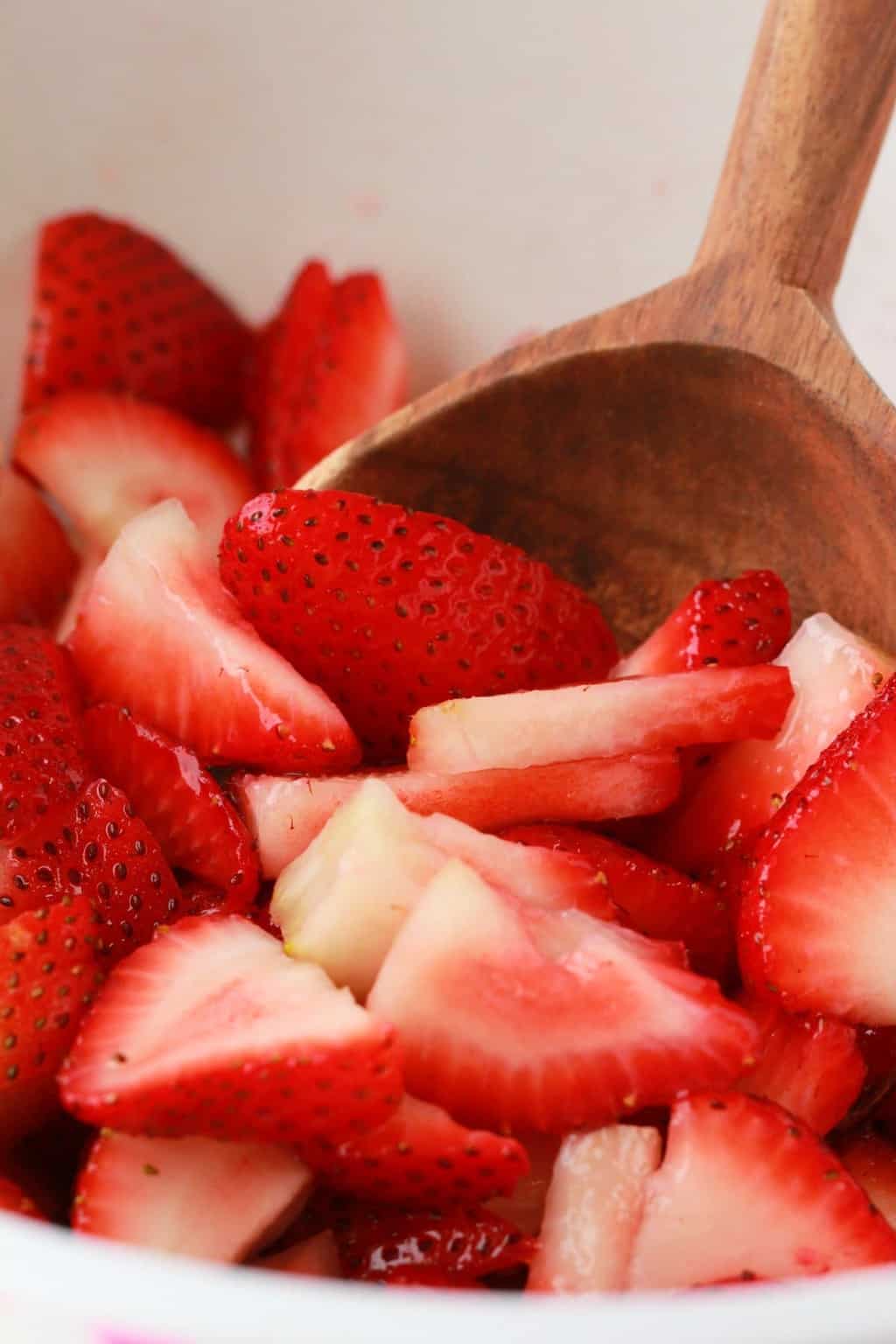Sliced strawberries in a mixing bowl with a wooden spoon. 