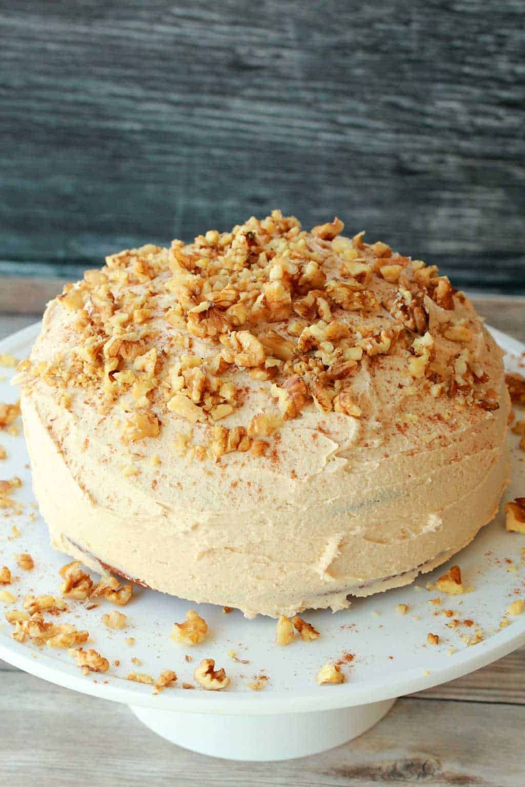 Vegan pumpkin cake topped with cinnamon buttercream frosting and crushed walnuts on a white cake stand. 
