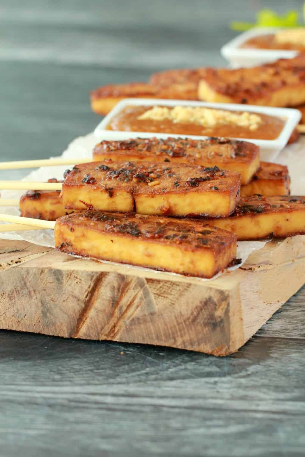 Tofu satay on a wooden board, peanut sauce in a white dish. 