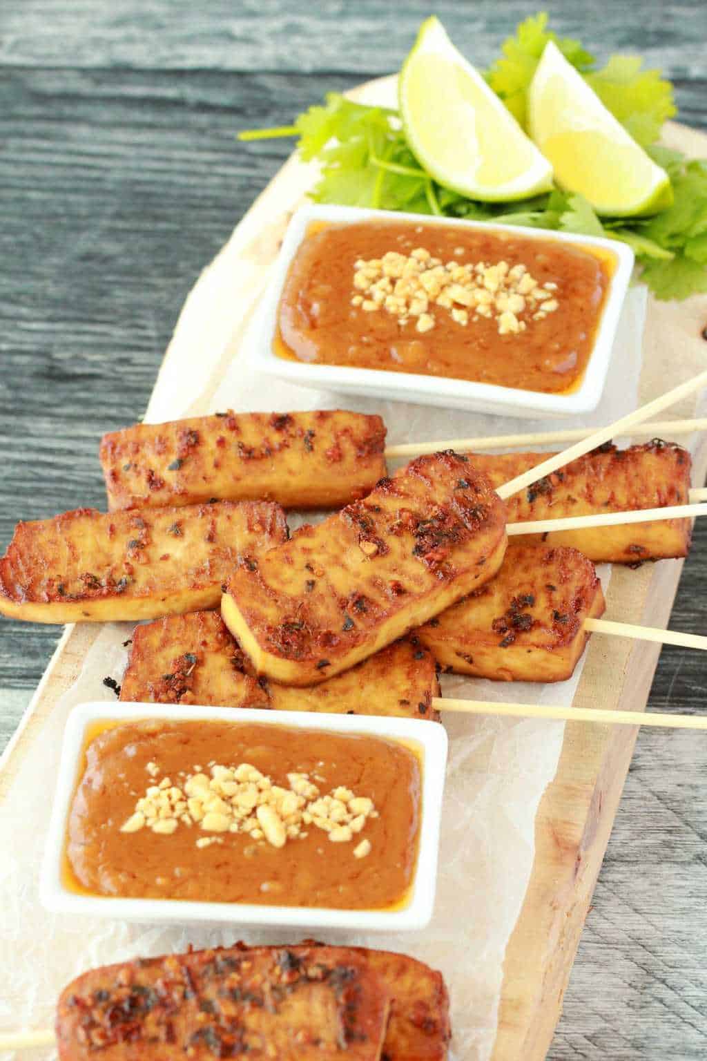 Tofu satay on a wooden board, peanut sauce in white dishes, cilantro and fresh lime. 