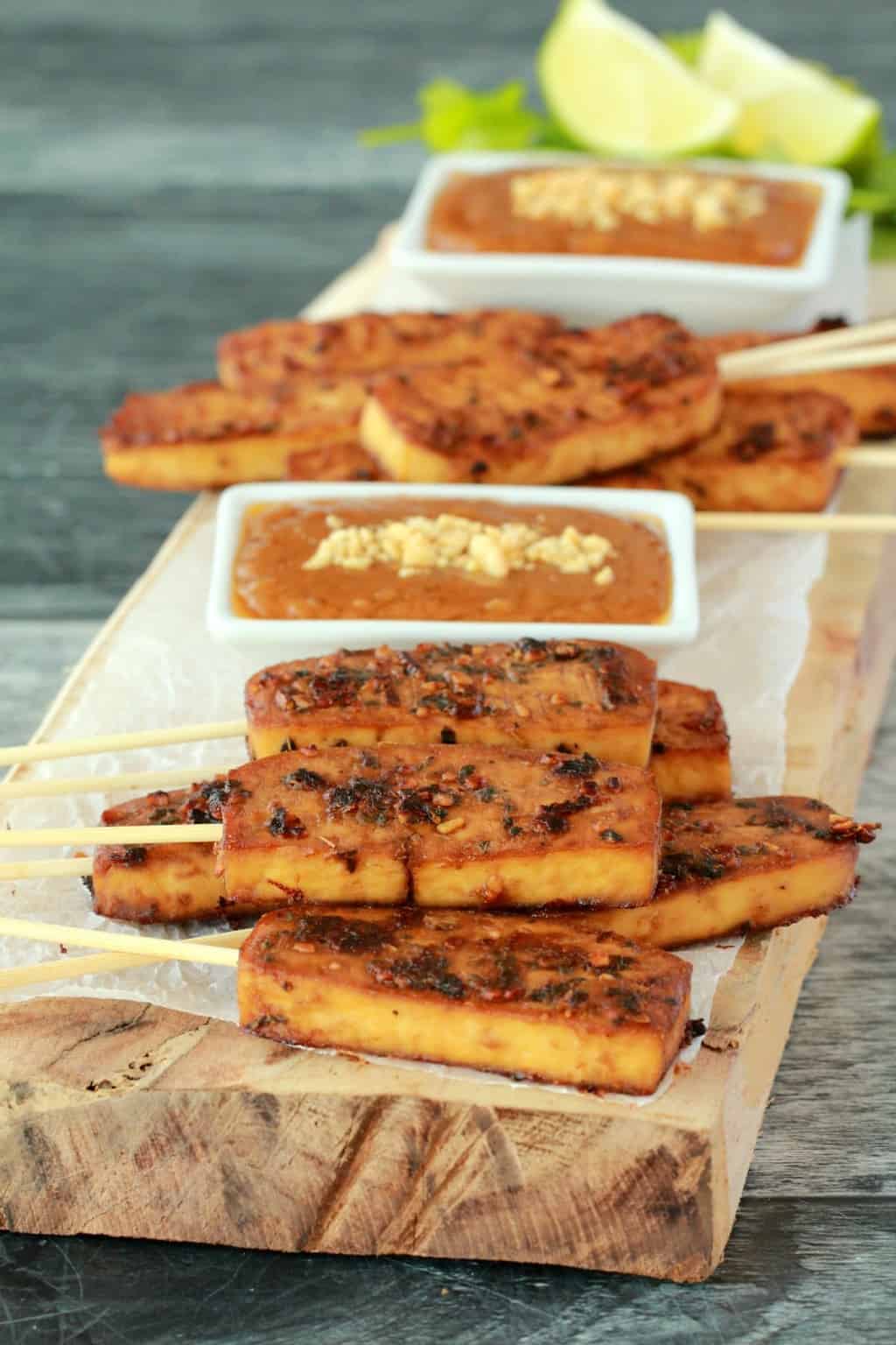 Baked tofu satay on a wooden board with white dishes of peanut sauce. 