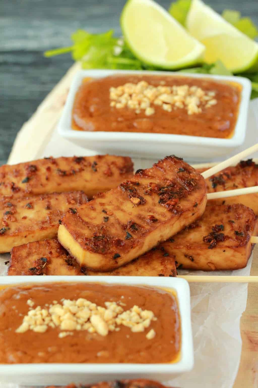 Baked tofu satay on a wooden board with white dishes of peanut sauce, cilantro and fresh lime. 