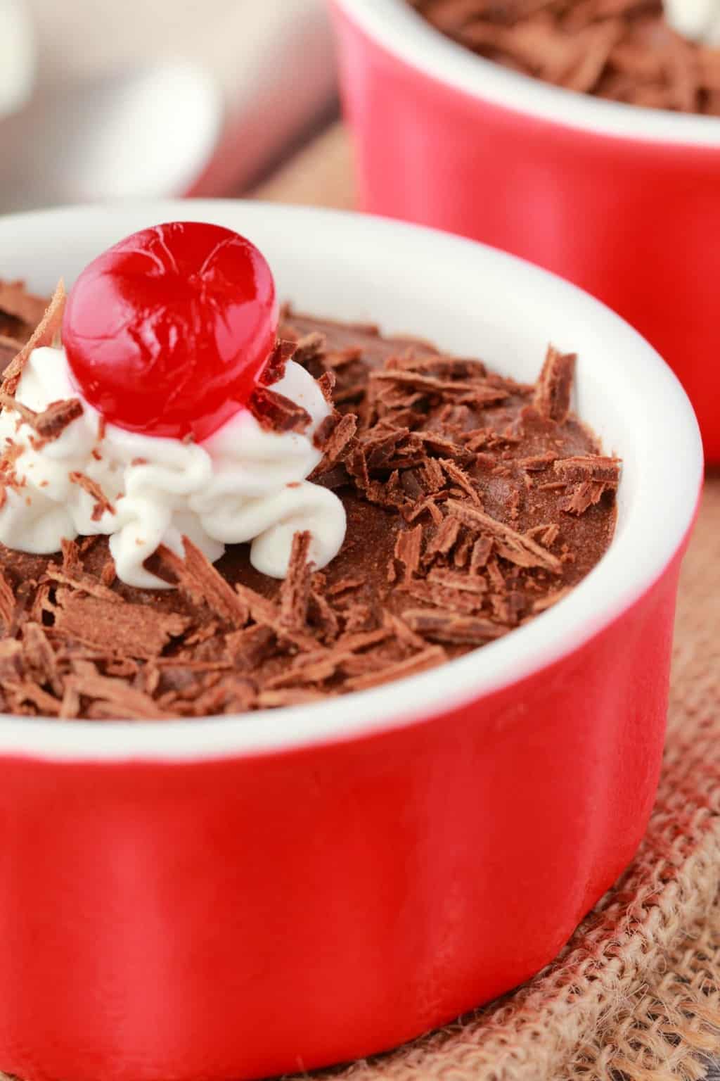 Vegan chocolate mousse in a red and white ramekin with chocolate shavings, a dollop of coconut cream and a cherry