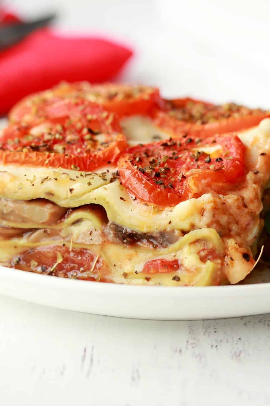 Slice of vegan lasagna topped with sliced tomato and black pepper on a white plate.