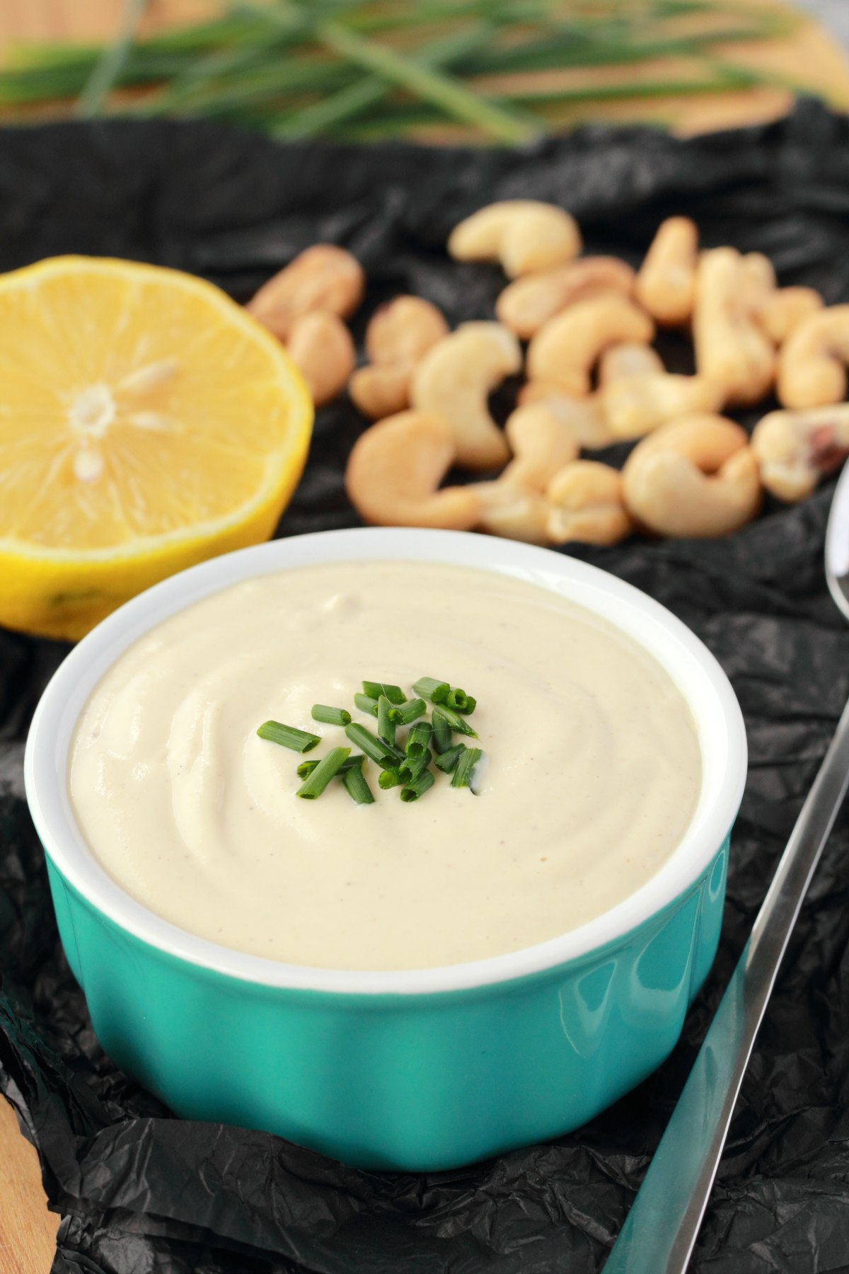 Cashew cream in a turquoise and white ramekin with chopped chives on top. Half a lemon, cashews and chives are in the background. 