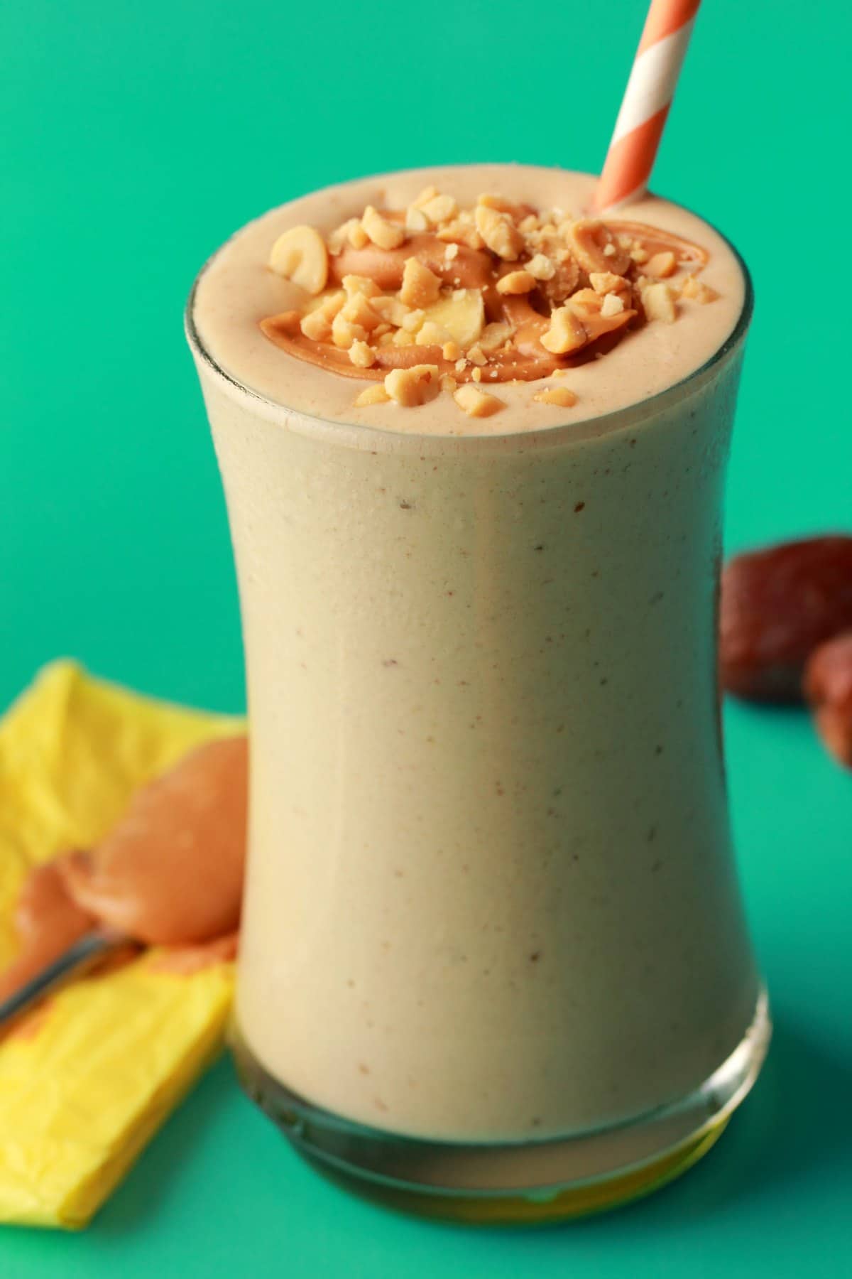 Peanut Butter Banana Smoothie in a glass with an orange and white straw. A spoonful of peanut butter and some medjool dates in the background. 
