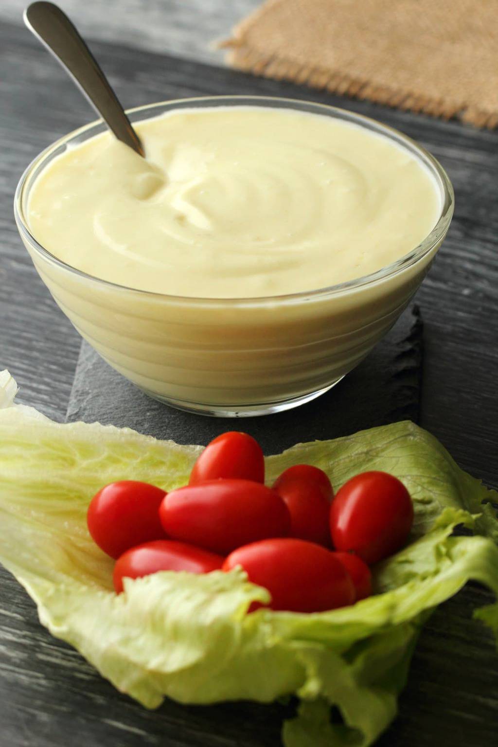 Vegan Mayonnaise in a glass bowl with a spoon and some cherry tomatoes and lettuce. 