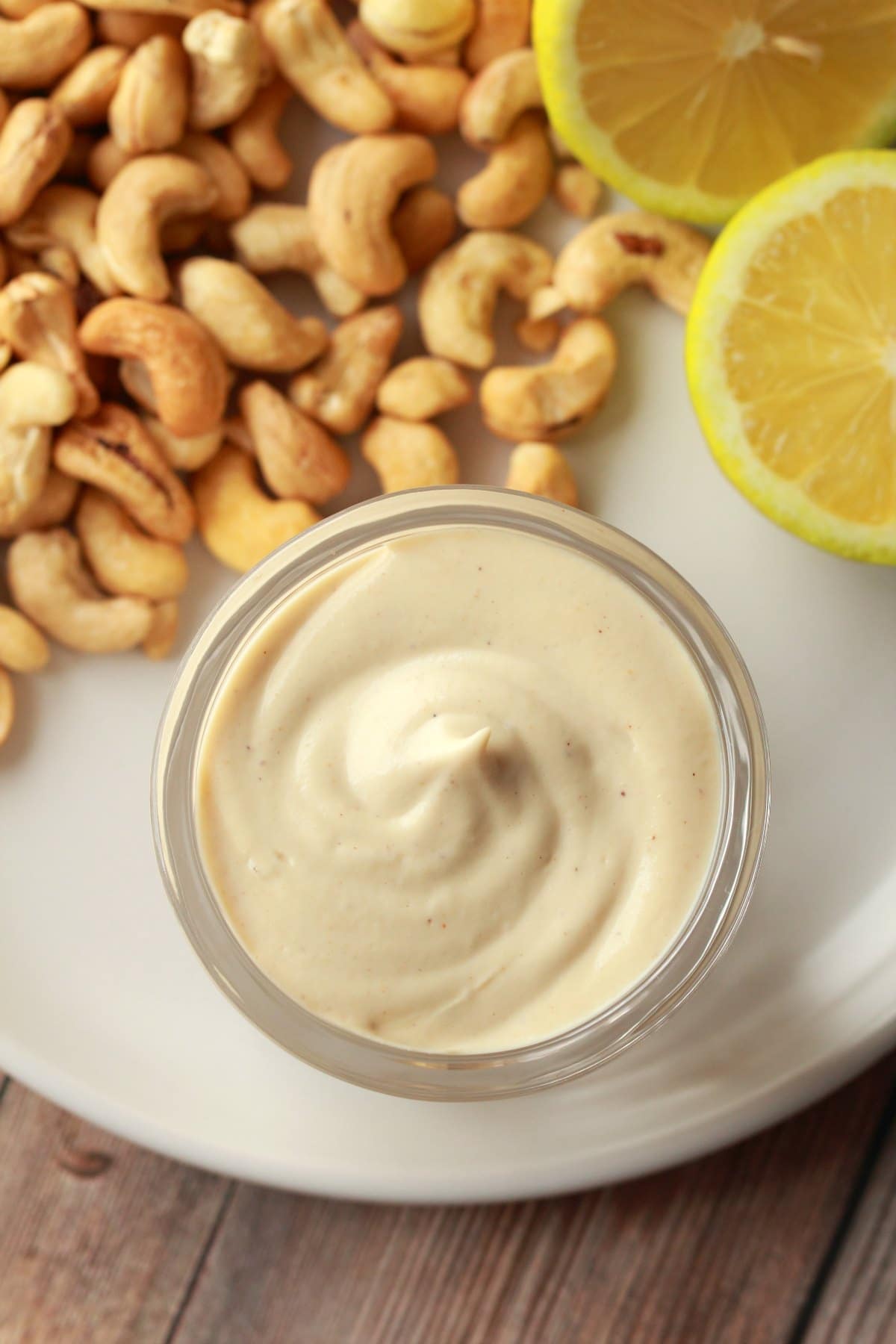 Vegan sour cream in a glass jar standing on a white plate with sliced lemon and cashews. 