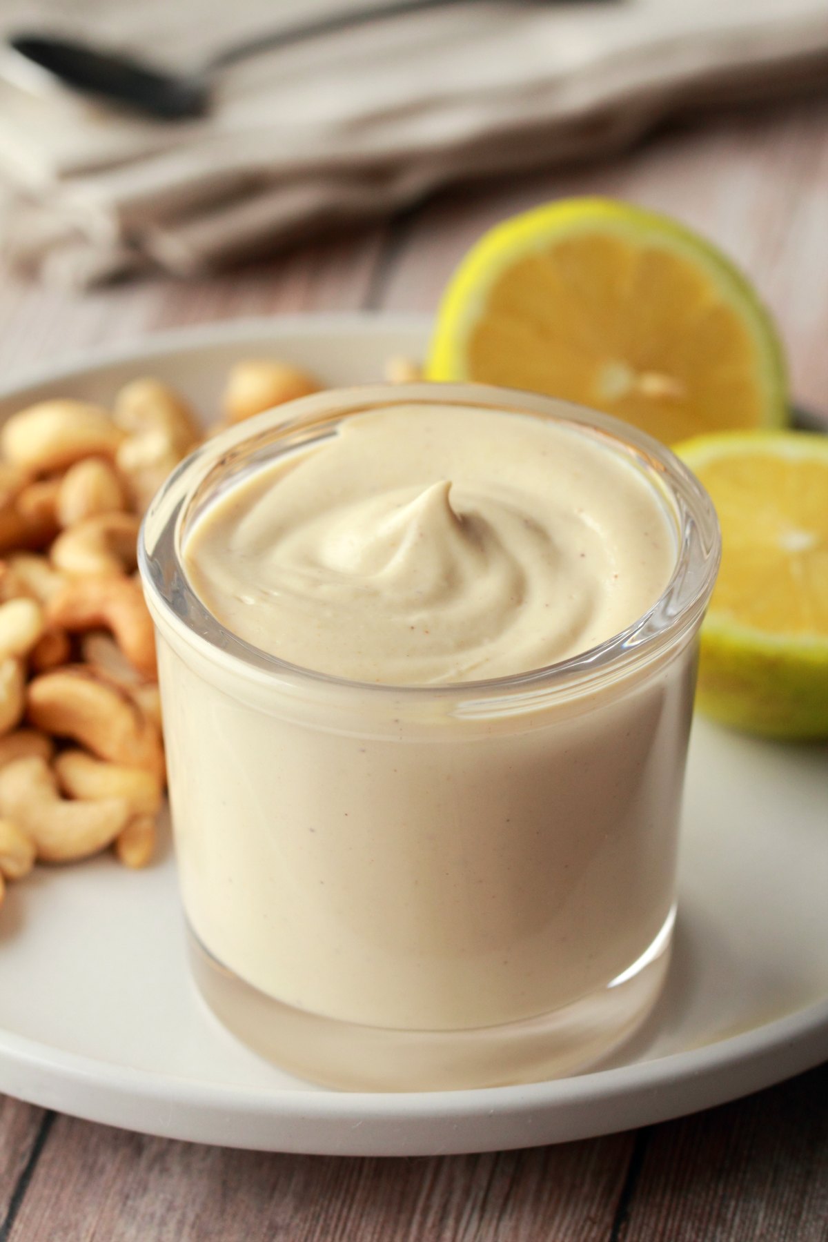 Vegan sour cream in a glass jar on a white plate with sliced lemon and cashews. 