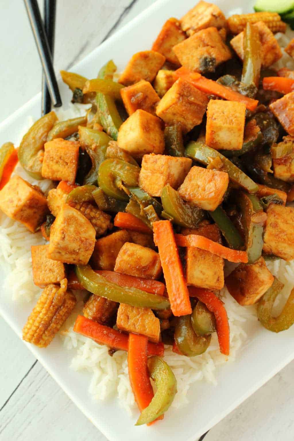 Tofu stir fry served over rice on a white plate with chopsticks. 