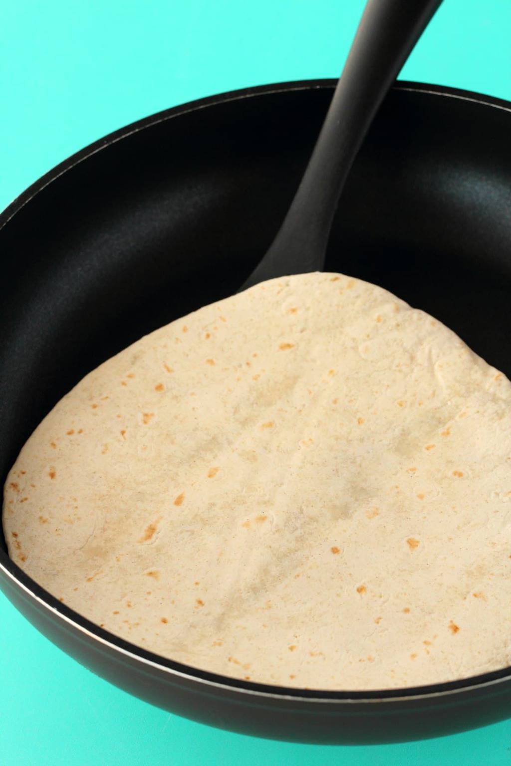 A flour tortilla in a frying pan with a spatula. 