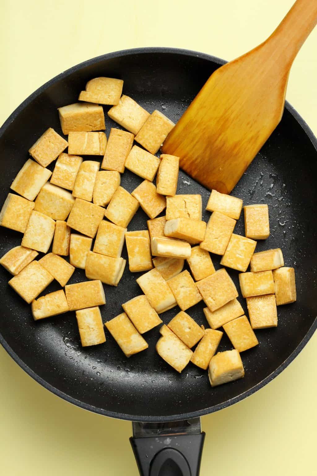 Cubes of fried tofu in a frying pan with a wooden spatula.