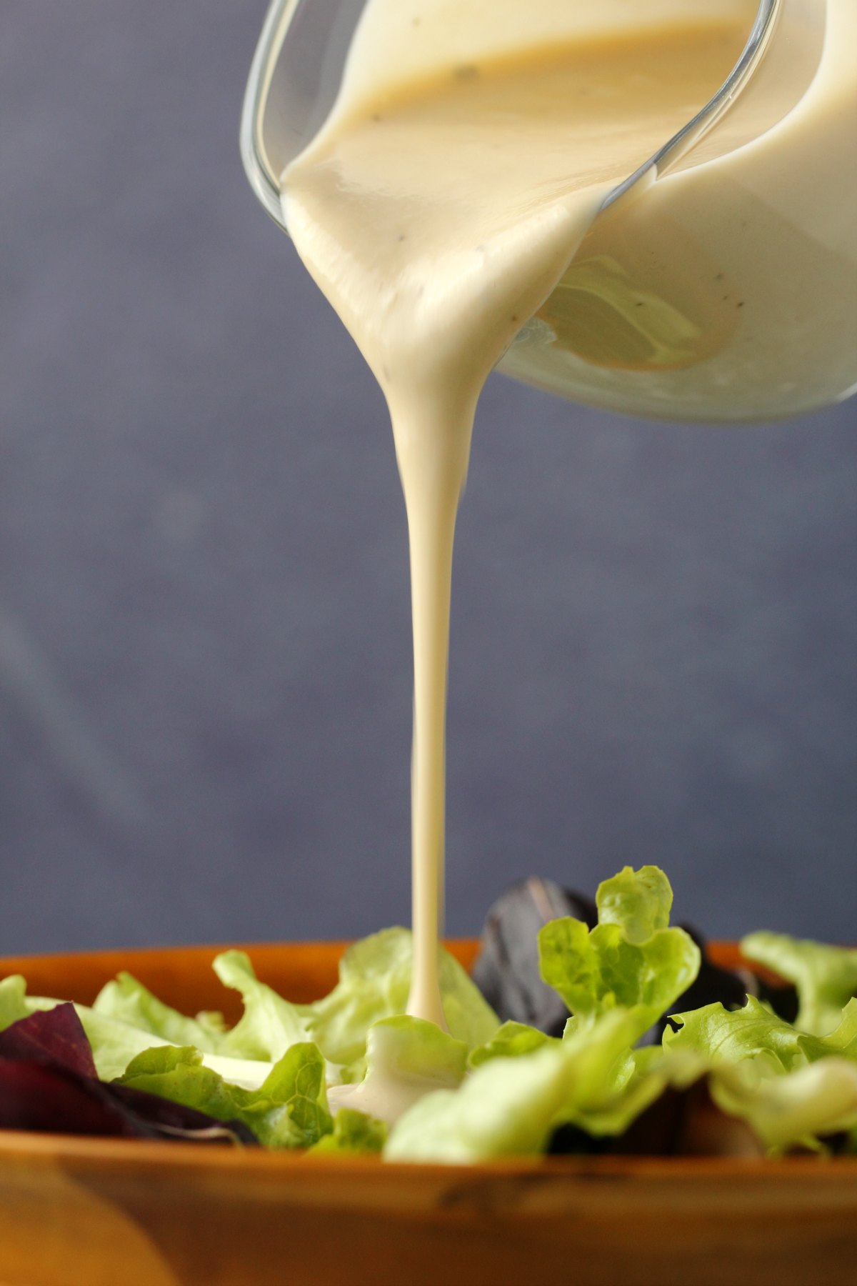 Tahini salad dressing pouring from a glass jug over salad greens in a bowl. 