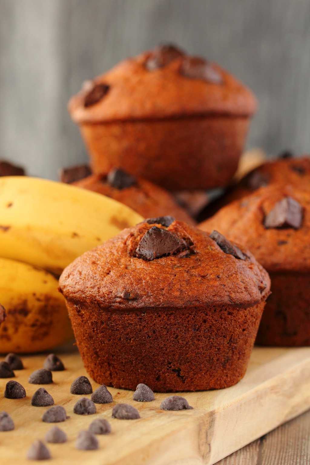 Vegan banana chocolate chip muffins on a wooden board with bananas and chocolate chips. 