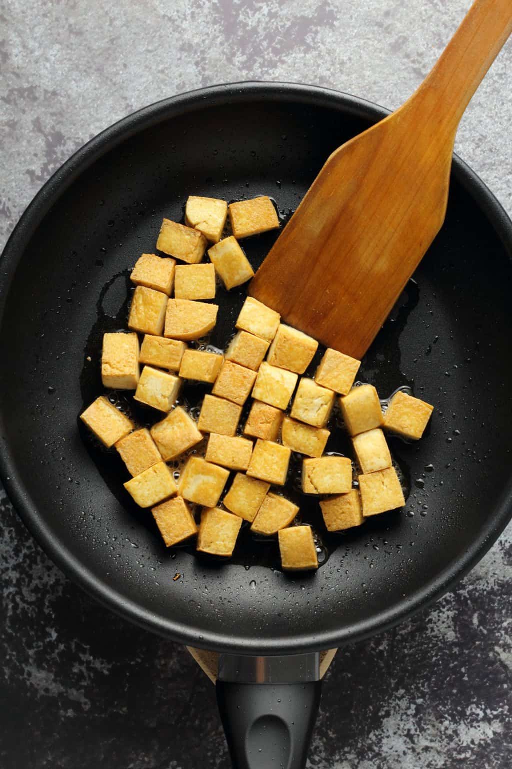 Cubes of tofu frying in a frying pan with a wooden spatula. 
