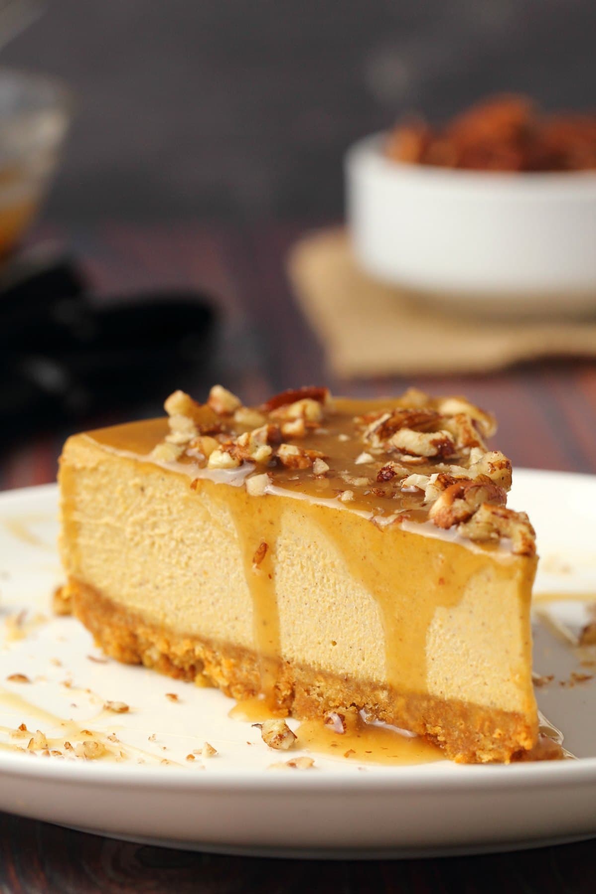 A slice of vegan pumpkin cheesecake topped with caramel sauce and crushed pecans on a white plate. 
