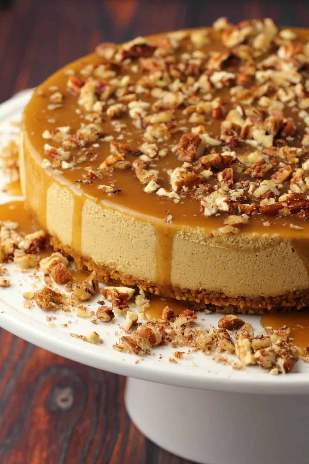 Vegan pumpkin cheesecake topped with caramel sauce and crushed pecans on a white cake stand. 