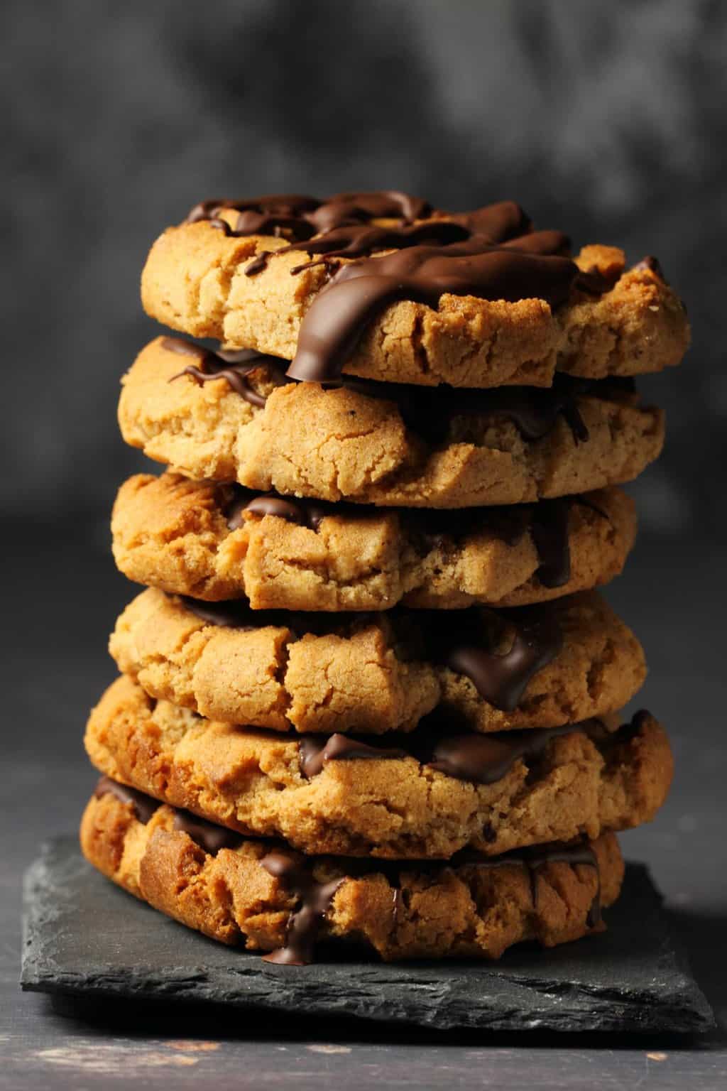 Vegan almond butter cookies topped with drizzled chocolate in a stack against a dark background. 