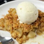 Slice of vegan apple crisp topped with a scoop of ice cream on a white plate.