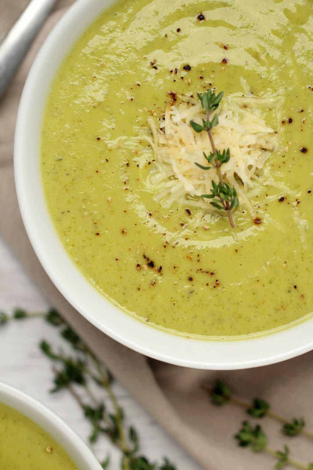 Vegan zucchini soup topped with vegan parmesan and a sprig of fresh thyme in a white bowl. 