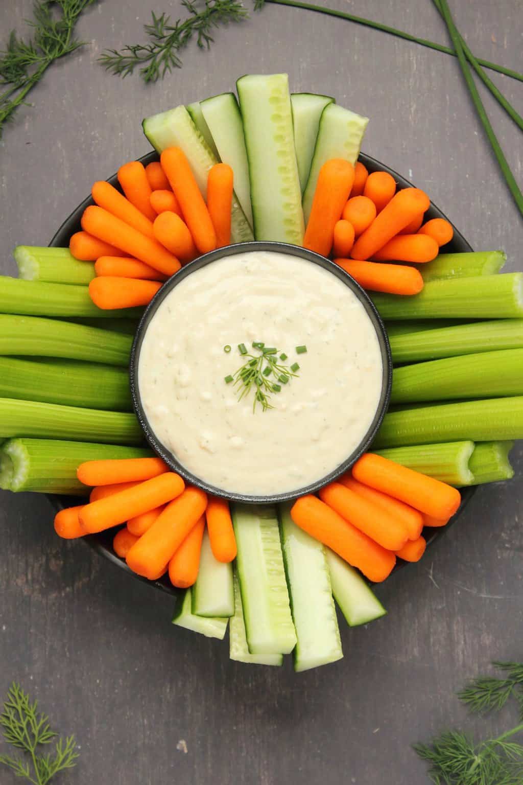 Vegan blue cheese dressing in a black bowl surrounded by celery, cucumber and baby carrots. 