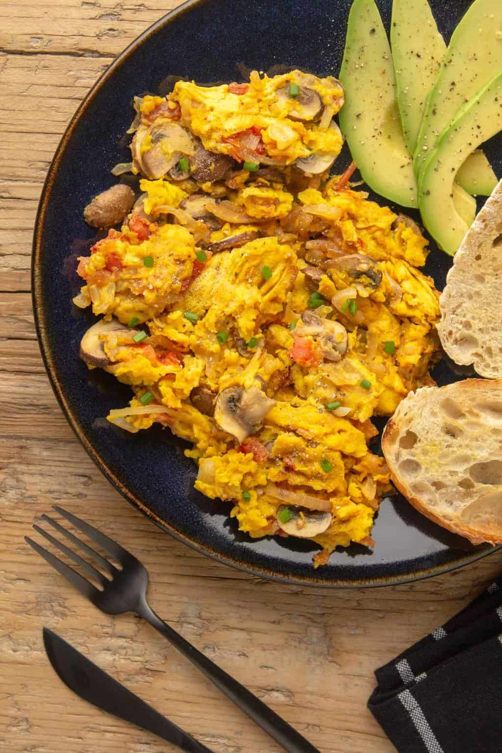 Vegan scrambled eggs with sliced avocado and toasted ciabatta on a black plate. 