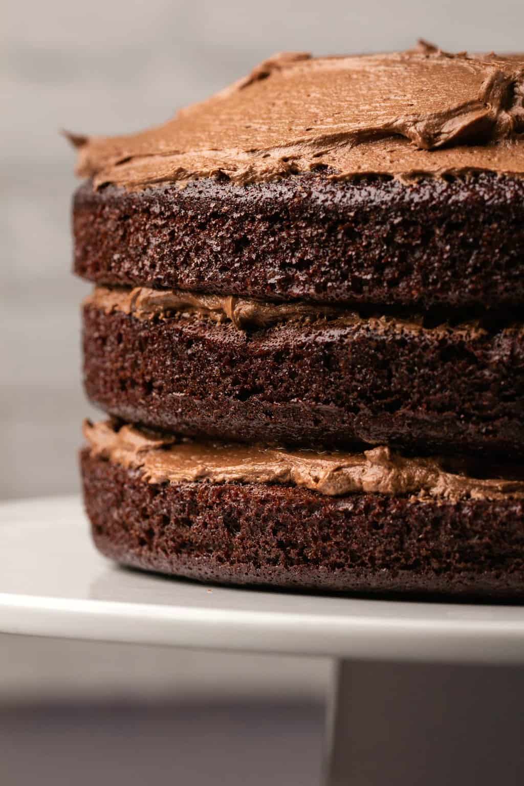 Three layer dairy free chocolate cake with naked sides on a white cake stand. 