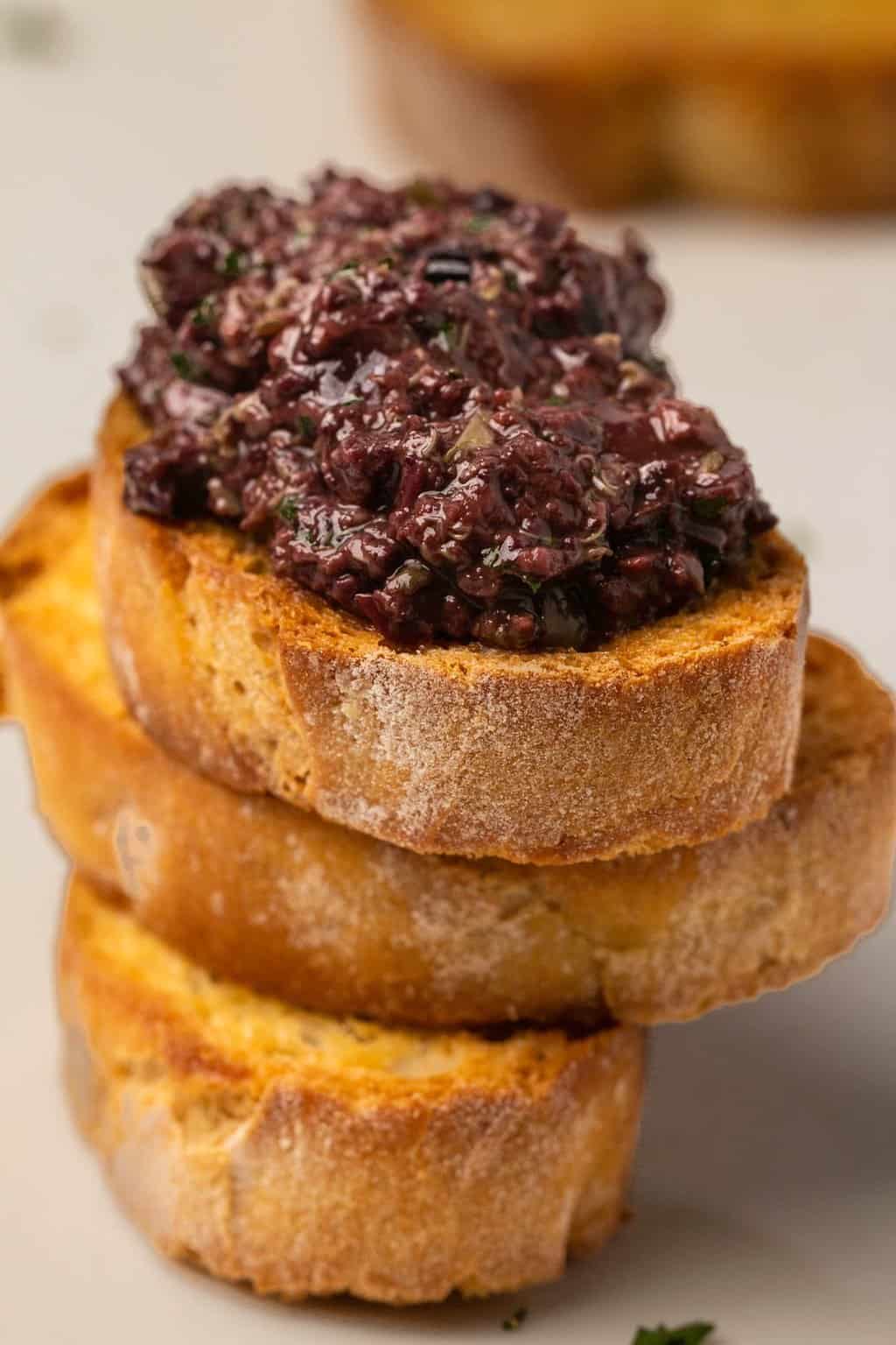Olive tapenade spread on top of a stack of toasted baguette slices. 