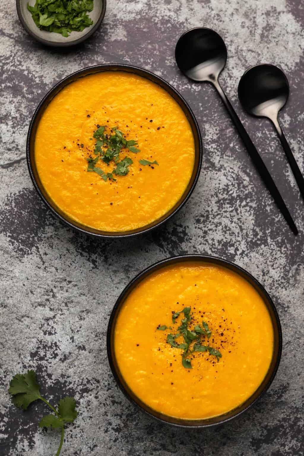 Vegan carrot ginger soup topped with chopped cilantro and black pepper in black bowls. 