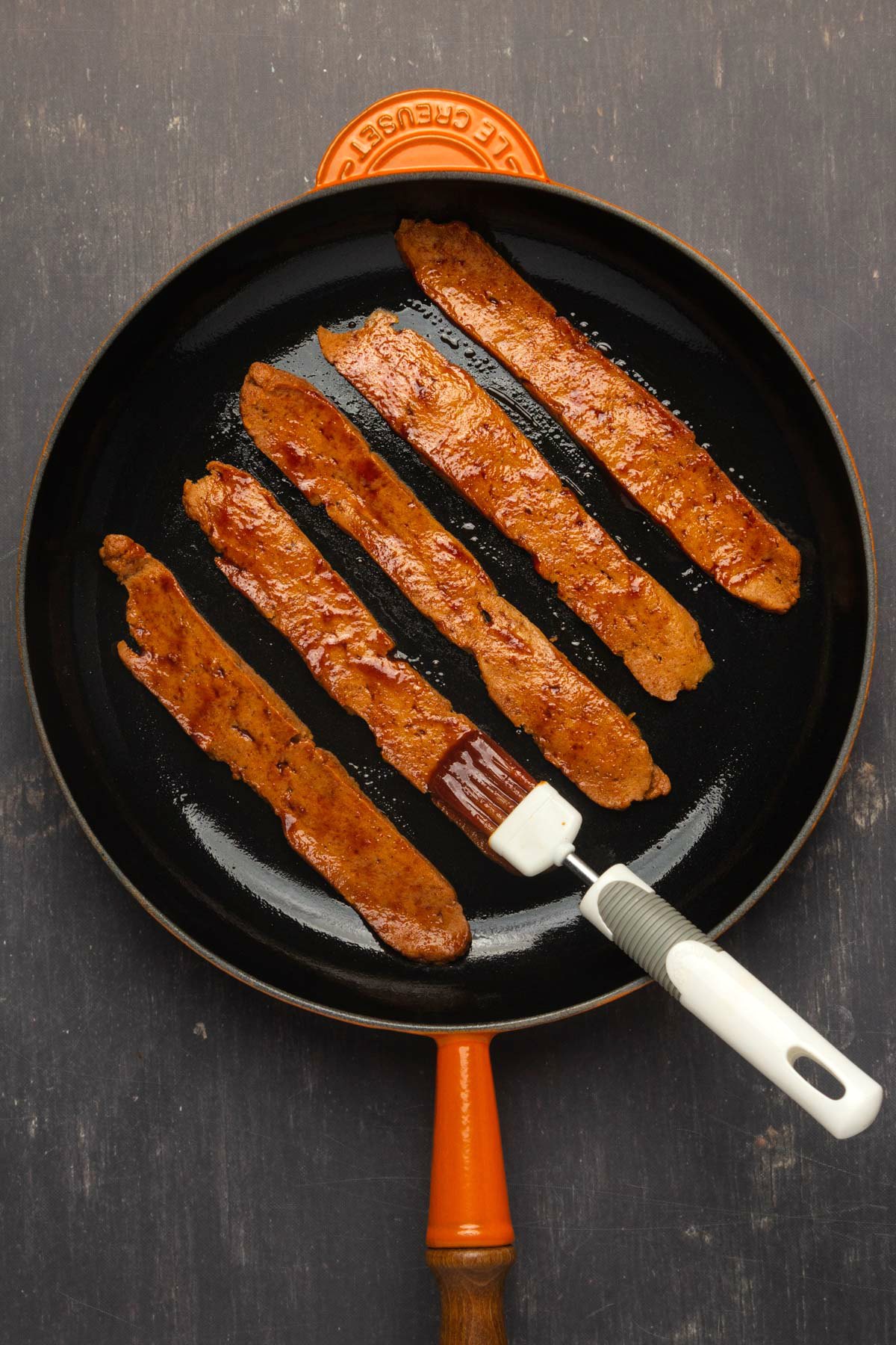 Vegan bacon strips in a frying pan with marinade brushed on top.