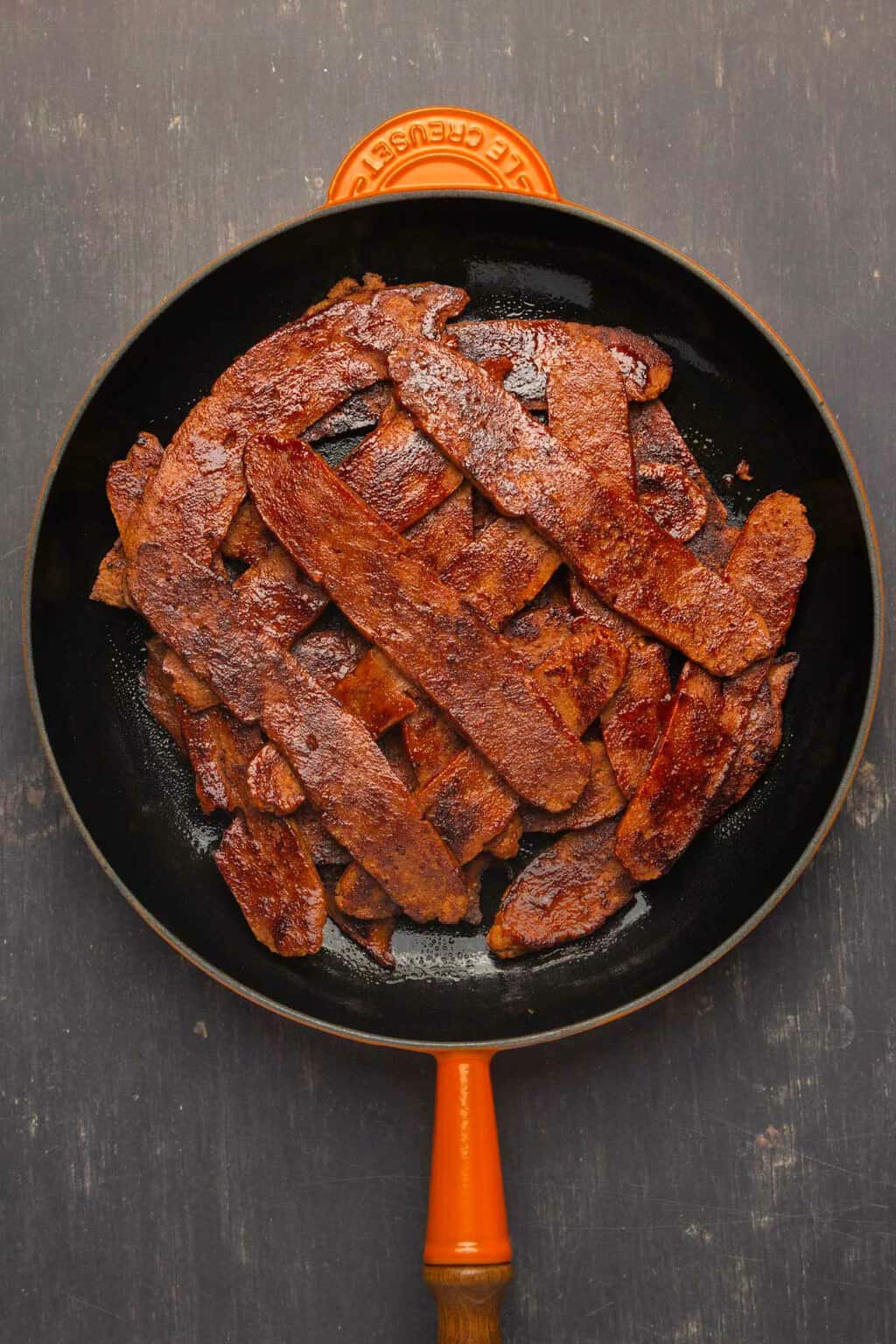 Vegan bacon strips stacked up in a frying pan. 