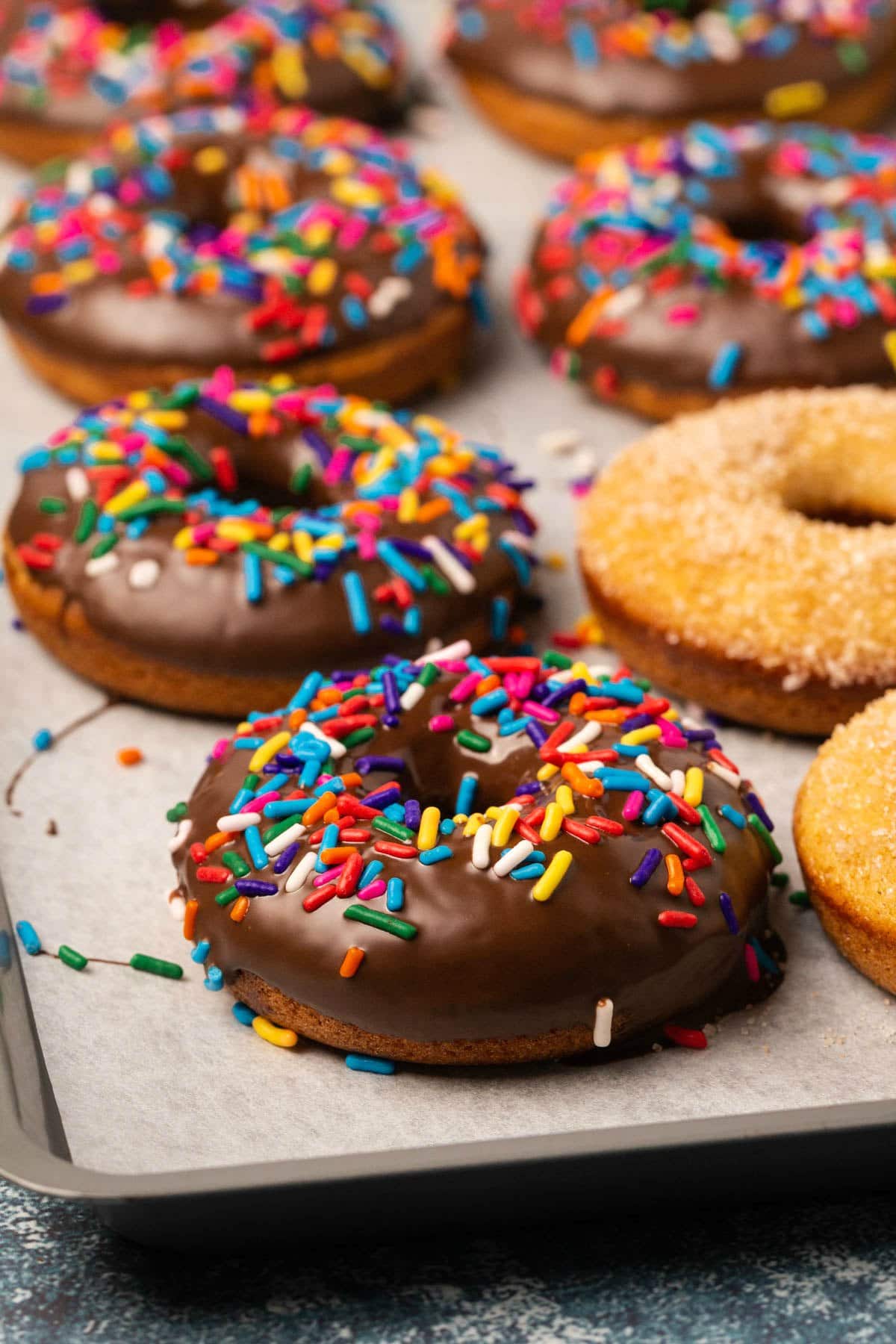 Vegan donuts topped with chocolate and sprinkles or cinnamon and sugar on a parchment lined baking sheet.