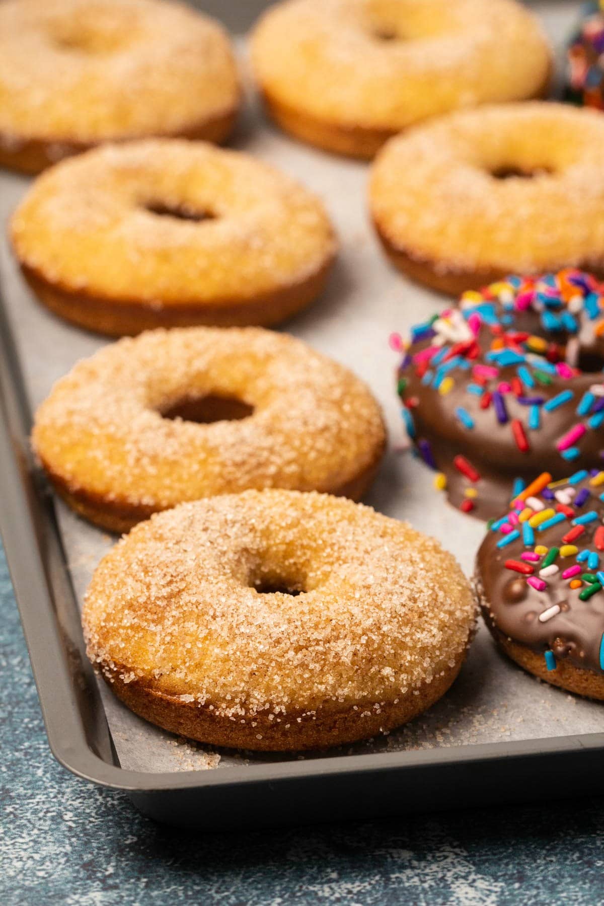 Vegan donuts topped with chocolate and sprinkles or cinnamon and sugar on a parchment lined baking sheet.