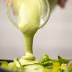 Cashew dressing pouring from a glass container onto a salad in a black bowl.
