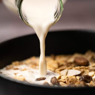 Almond milk pouring from a glass milk bottle onto a bowl of cereal.