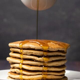 Syrup pouring from a glass jug over a stack of vegan protein pancakes on a white plate.