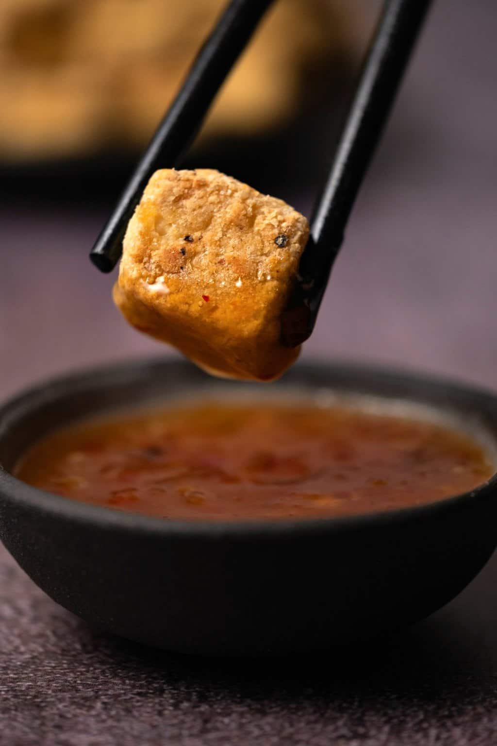 A cube of baked tofu dipping into a bowl of sweet chili sauce. 