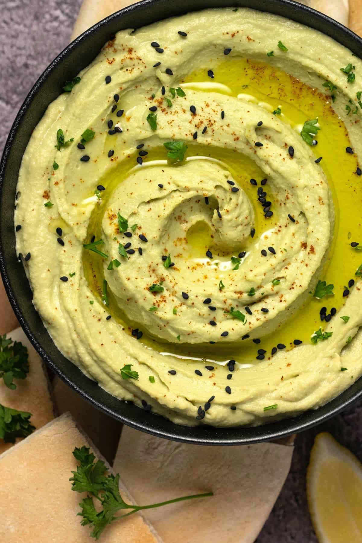 Avocado hummus in a black bowl with sesame seeds, olive oil and chopped parsley. 