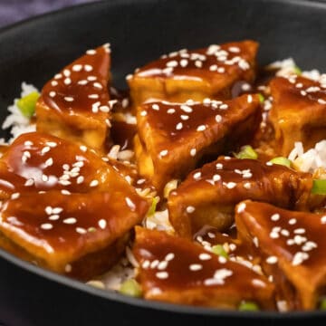 Teriyaki tofu with rice, sesame seeds and green onions in a black bowl.