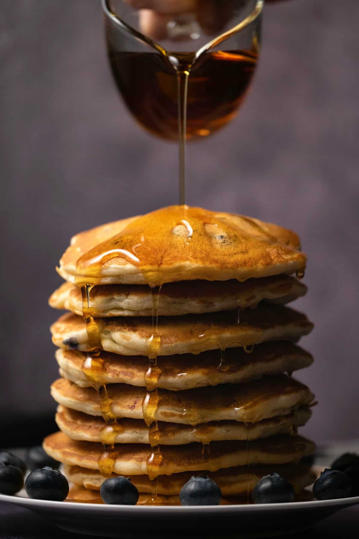 Syrup pouring from a glass jug over a stack of pancakes. 