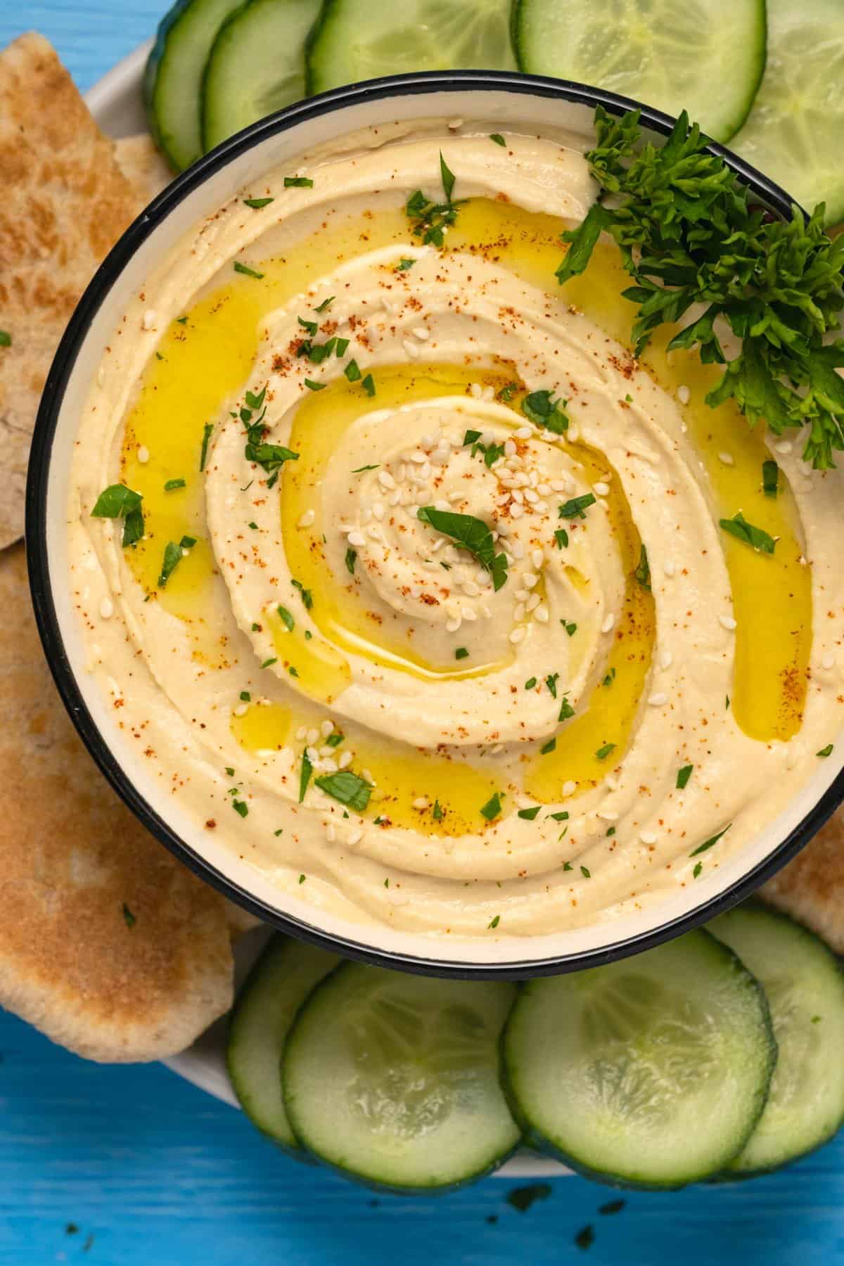 Hummus in a ceramic bowl with pita breads and sliced cucumber on the side. 