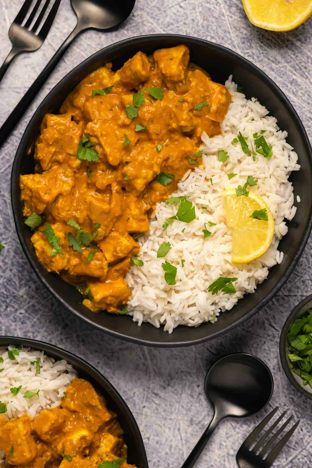 Vegan butter chicken in a black bowl with basmati rice, chopped cilantro and a slice of lemon. 