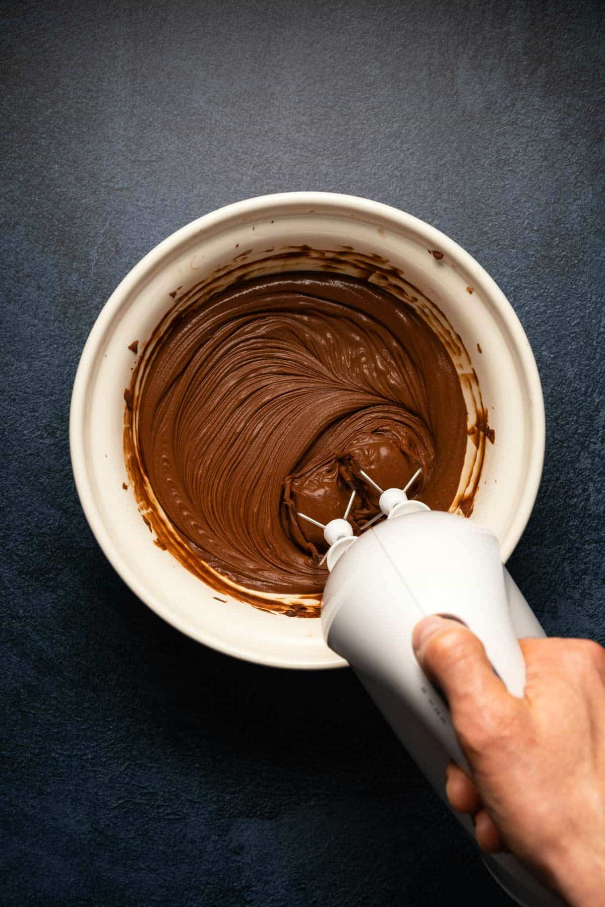 Whipping chocolate ganache with an electric whisk in a beige mixing bowl. 
